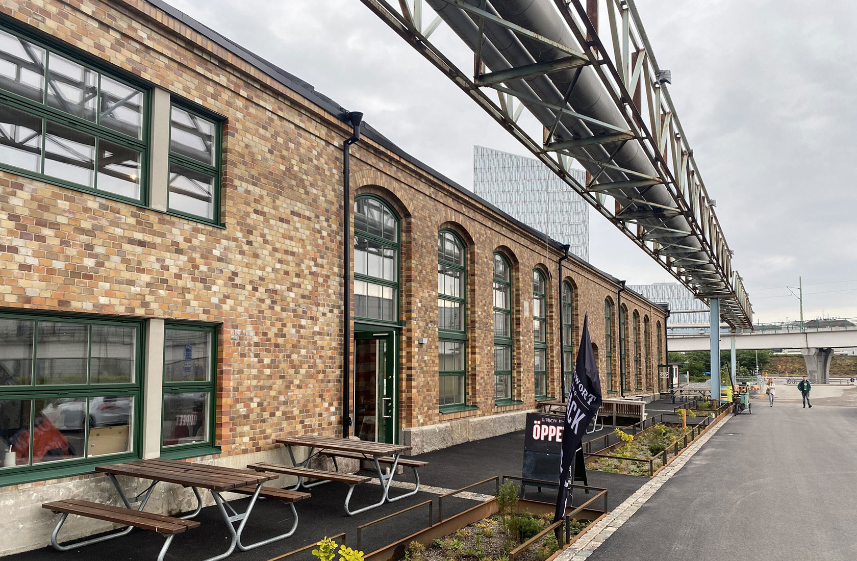 Old brick industrial building with green-framed windows, outside seating, and overhead pipes in urban setting.