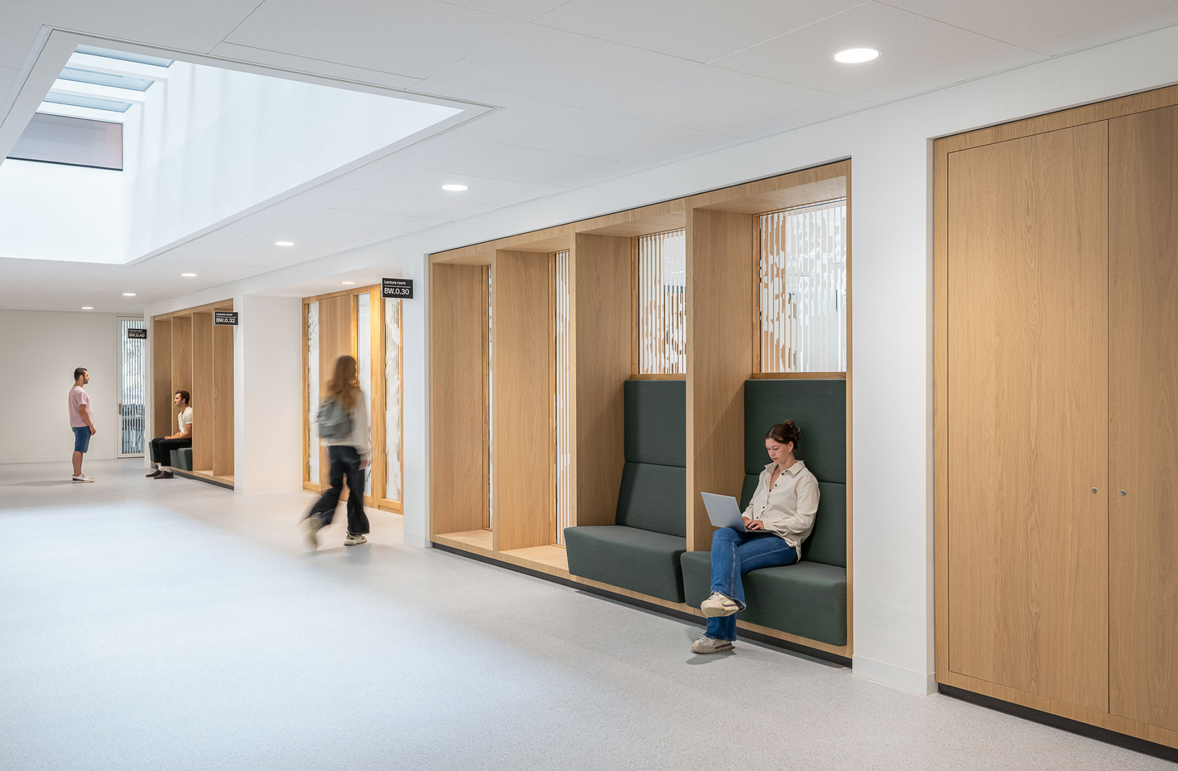 Modern hallway with built-in wooden seating, person using laptop, and walking people under skylight.