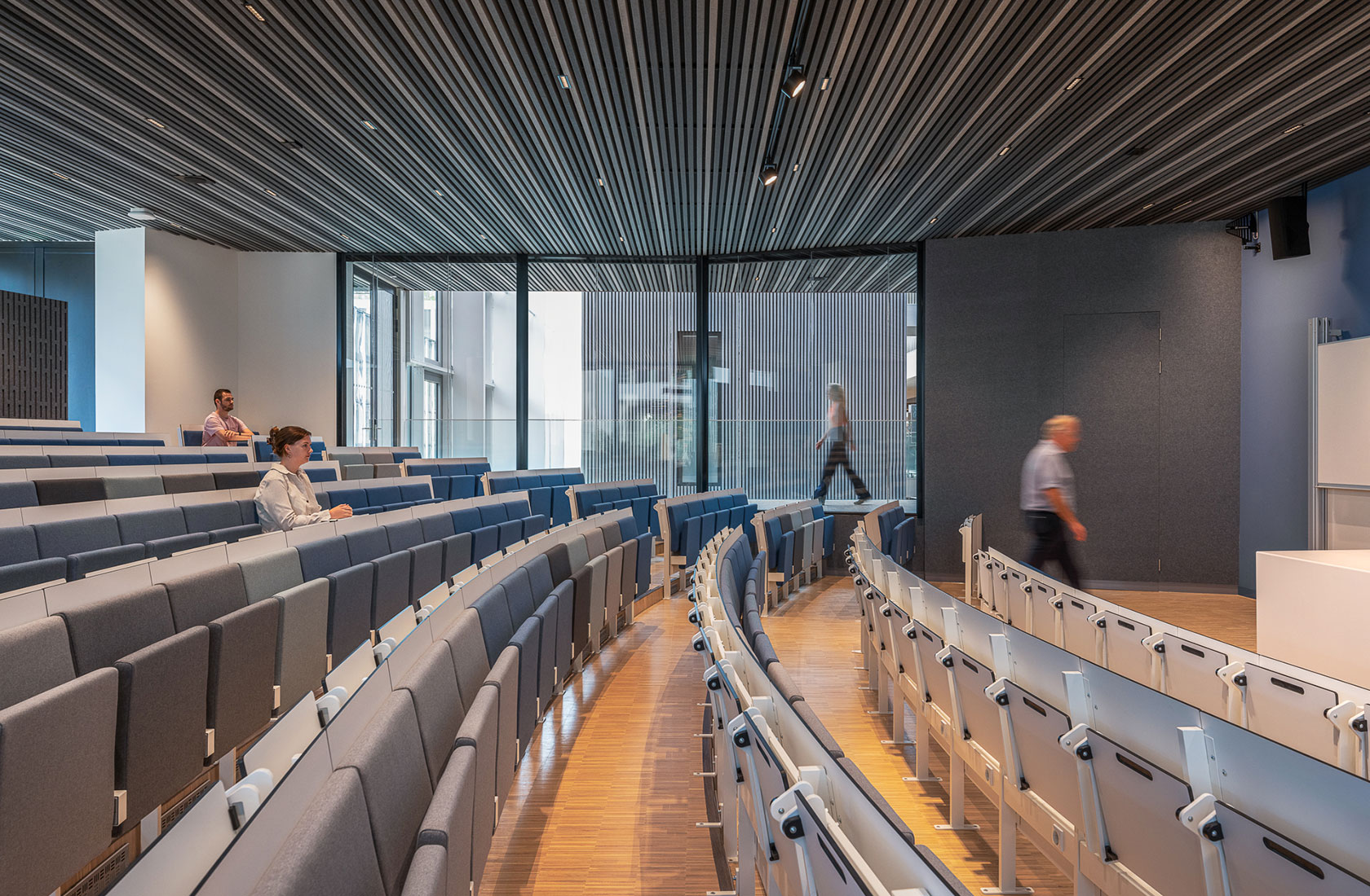 Modern lecture hall with tiered seating and a few people, featuring large windows and wooden flooring.
