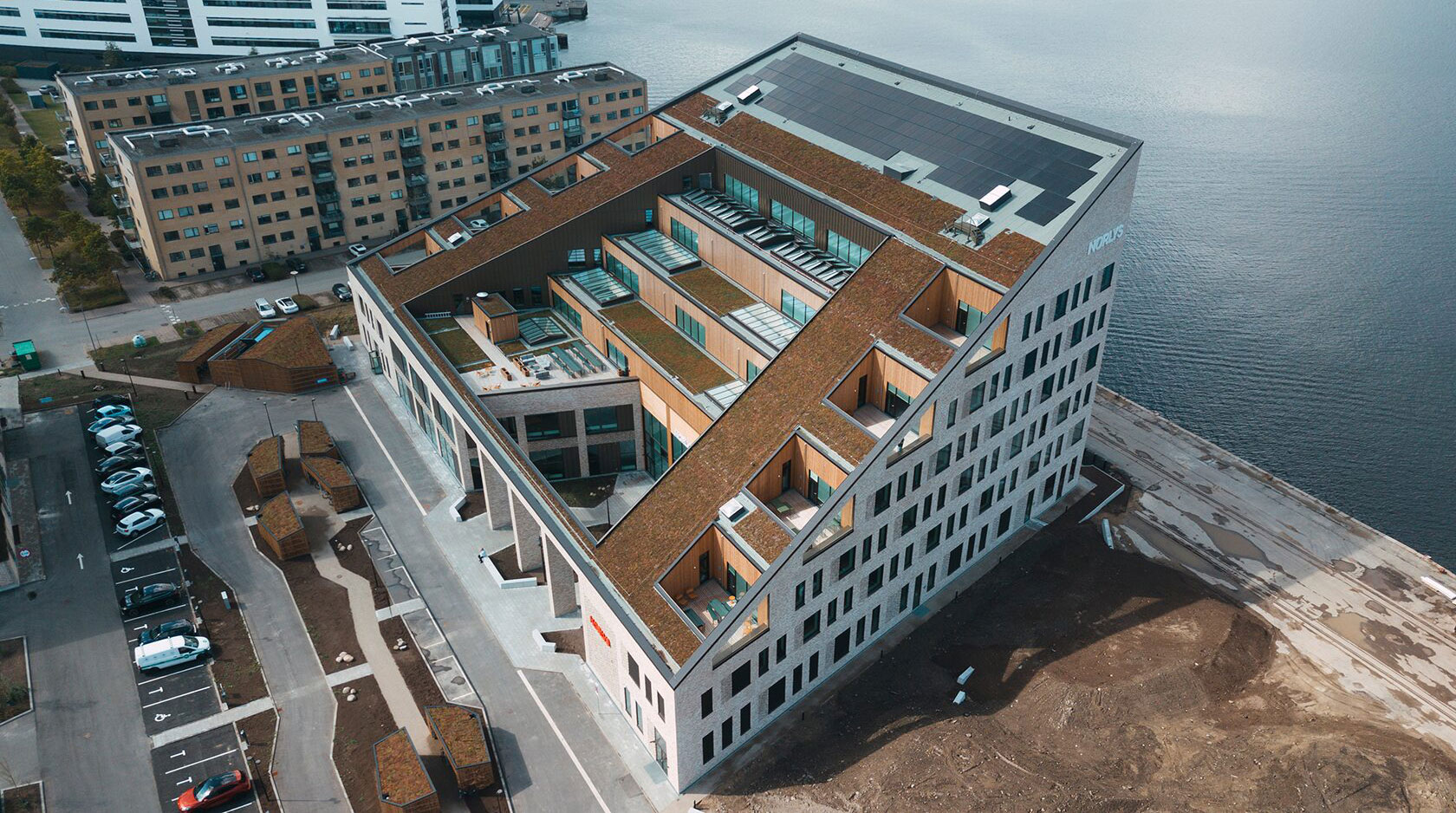 Modern waterfront building with green rooftop, surrounded by parking and adjacent to other buildings.