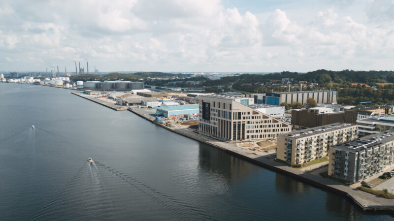 Aerial view of harbor with modern buildings, industrial tanks, and boats on a sunny day.