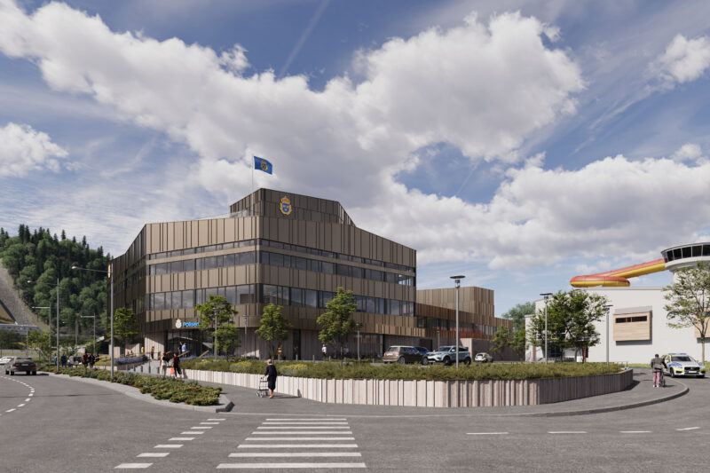 Modern police station building with flag, cars, and pedestrians in front of a mountain under cloudy sky.