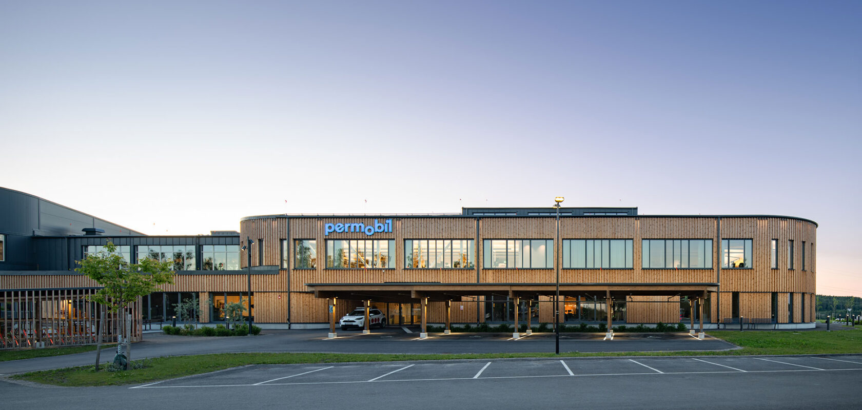 Modern office building with "permobil" signage, large windows, and wooden exterior at sunset.