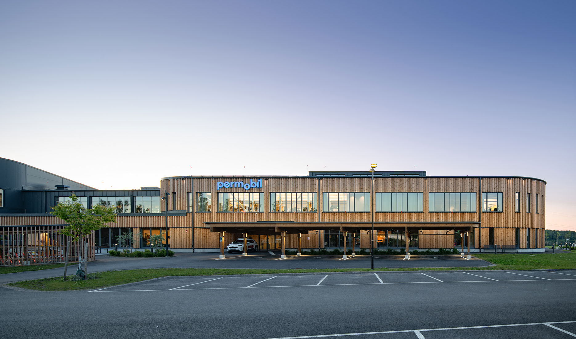 Modern office building with "permobil" signage, large windows, and wooden exterior at sunset.