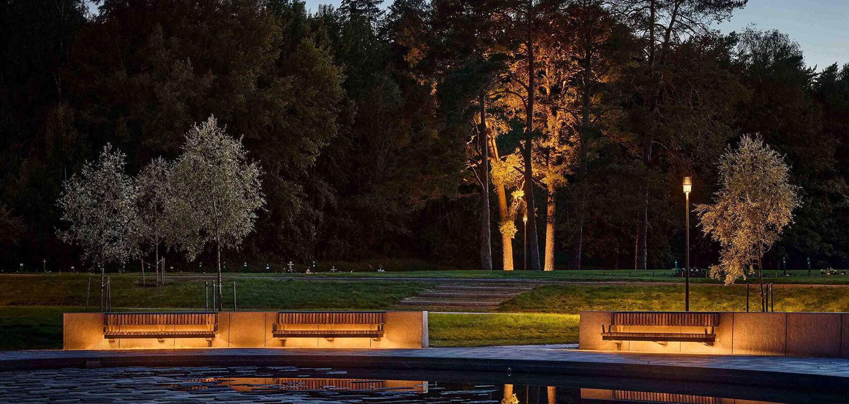 Cementery park at twilight with illuminated benches, reflecting pool, and trees in the background.