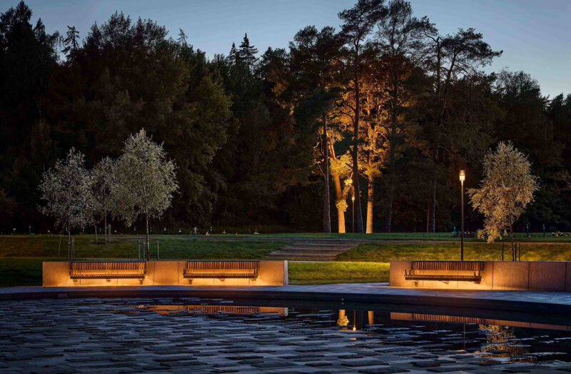 Cementery park at twilight with illuminated benches, reflecting pool, and trees in the background.