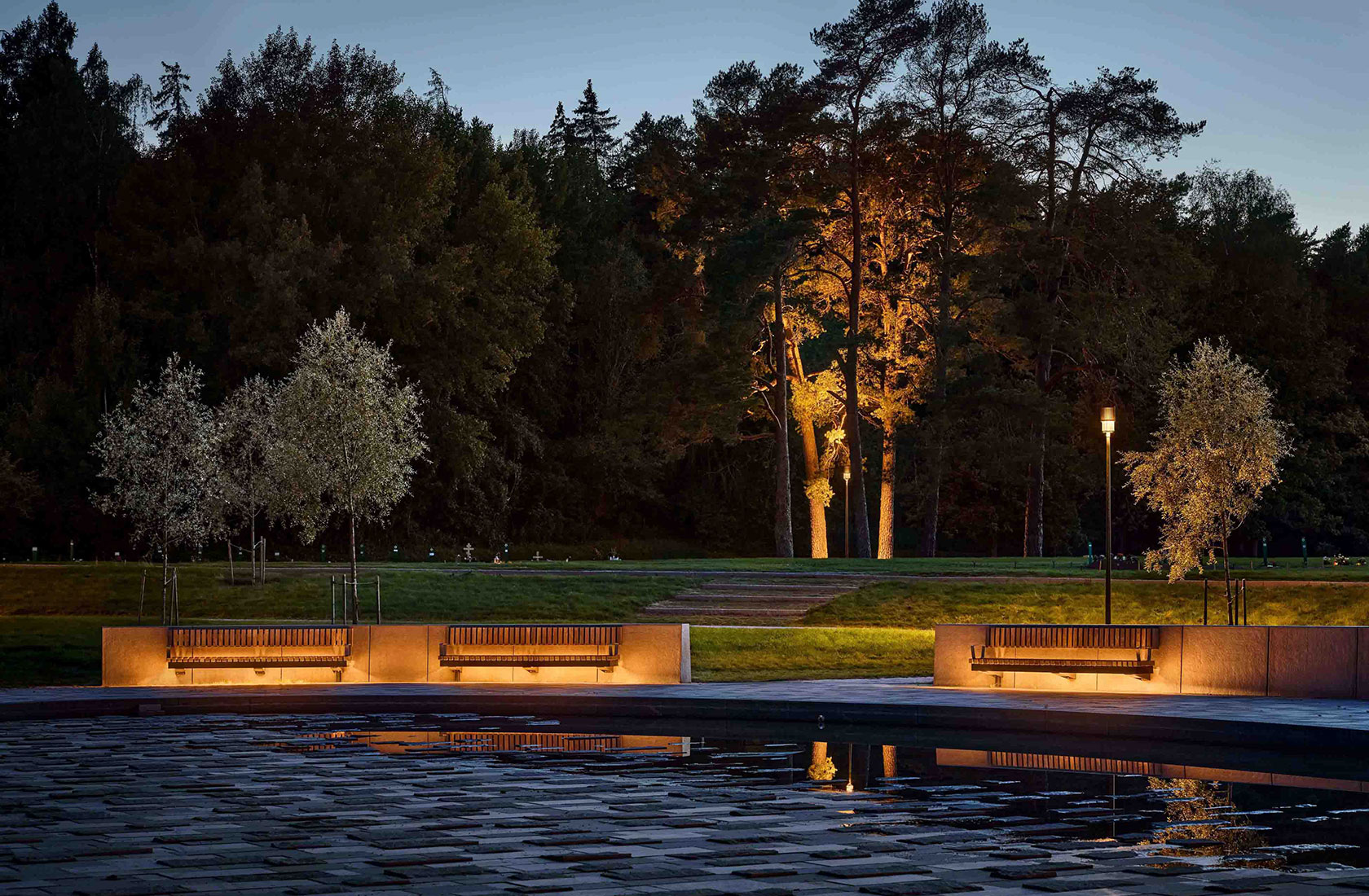 Cementery park at twilight with illuminated benches, reflecting pool, and trees in the background.