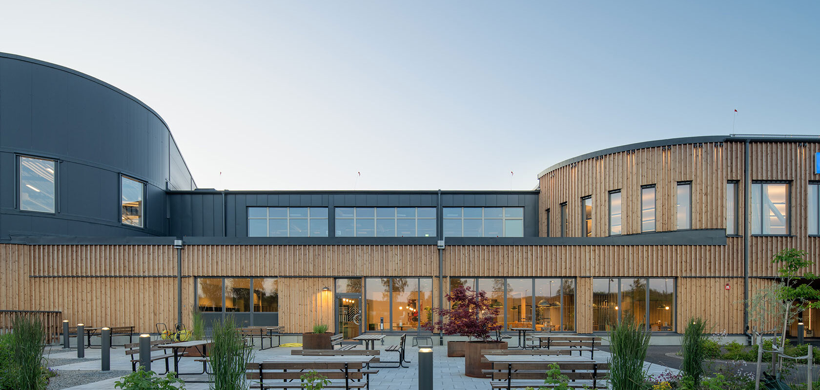 Modern building with large windows and wooden facade, featuring outdoor seating areas and landscaped gardens.