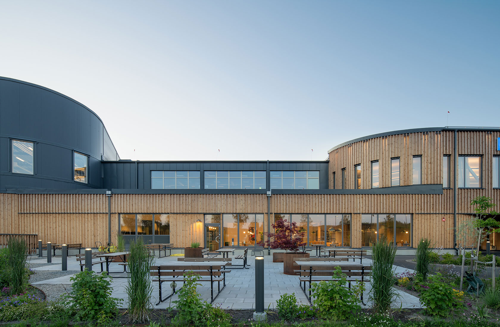 Modern building with large windows and wooden facade, featuring outdoor seating areas and landscaped gardens.