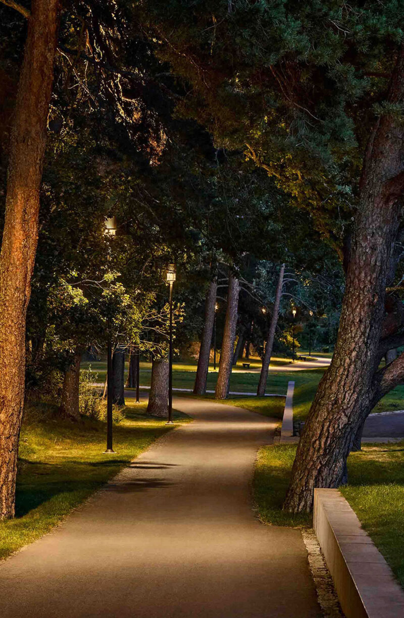 A well-lit winding pathway through a cementery in the evening, illuminated by street lamps.
