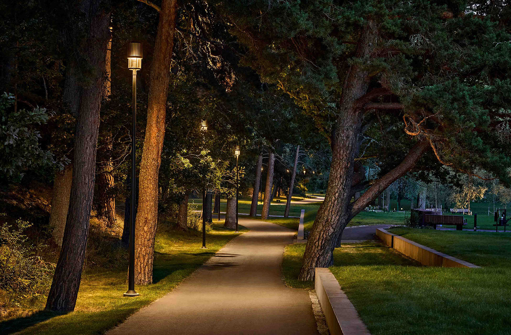 Cementery park pathway illuminated by street lamps at dusk, surrounded by trees and greenery.