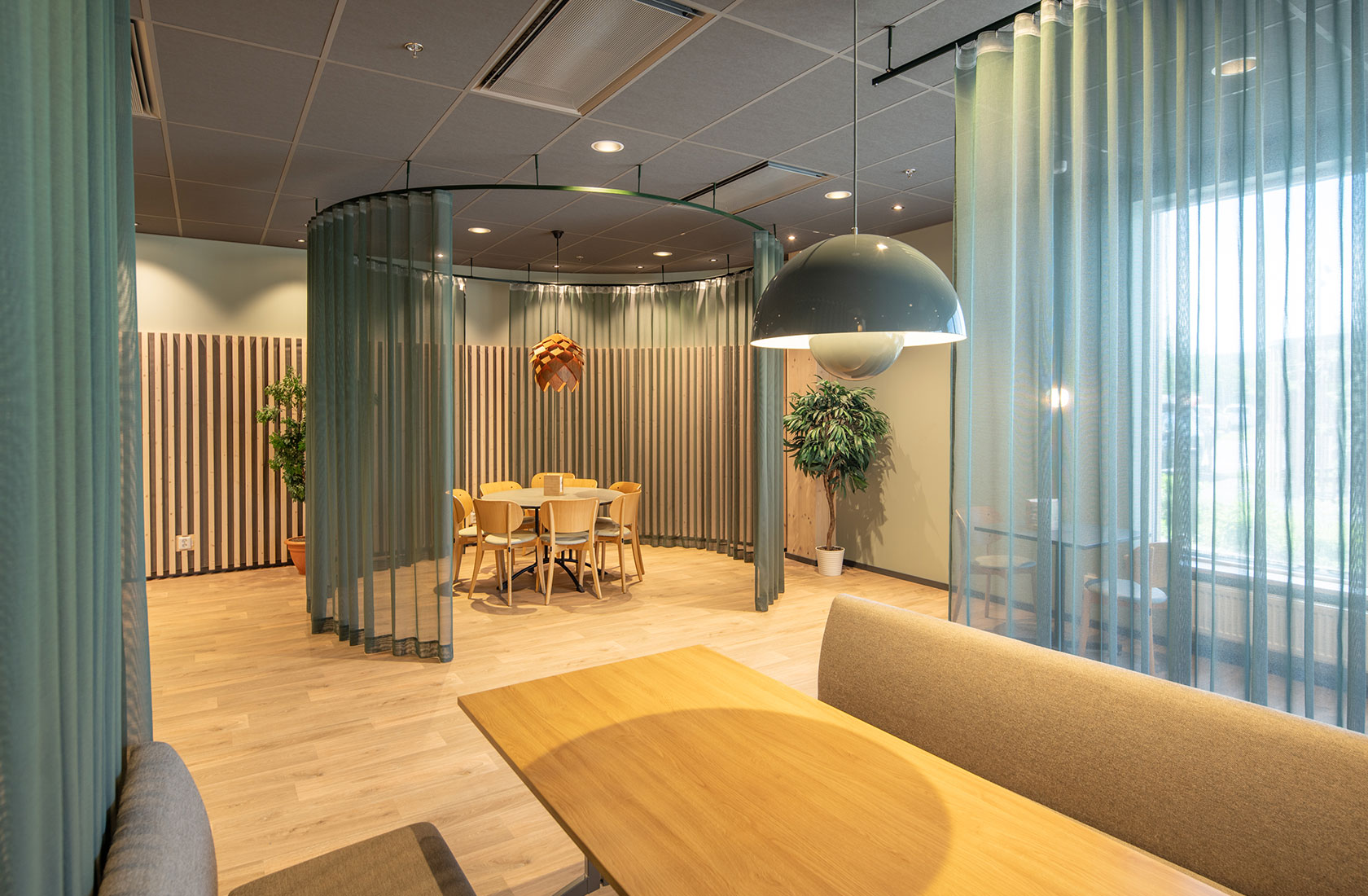 Modern office meeting space with wooden tables, chairs, green curtains, plants, and bright lighting.