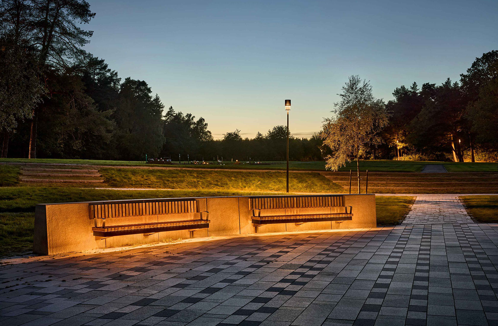 Park benches illuminated at sunset with trees in the background and paved walkway in foreground.