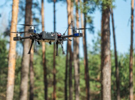 A drone hovers steadily above a lush forest, surrounded by tall trees. Its rotors spin softly as it captures stunning aerial shots of the vibrant greenery below on a clear day.