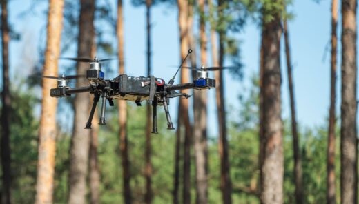 A drone hovers steadily above a lush forest, surrounded by tall trees. Its rotors spin softly as it captures stunning aerial shots of the vibrant greenery below on a clear day.