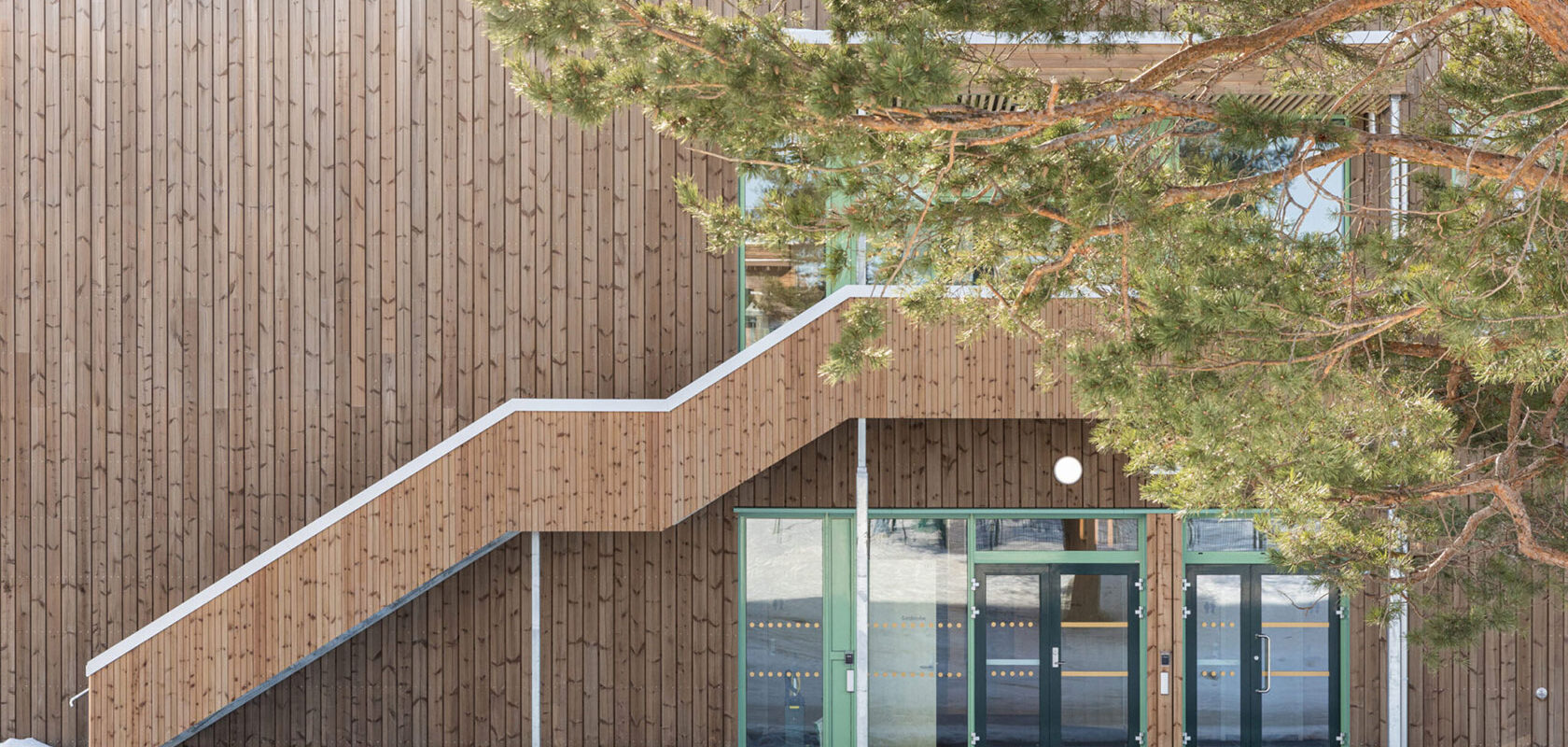 Modern wooden building with green framed glass doors and staircase amidst snowy landscape.