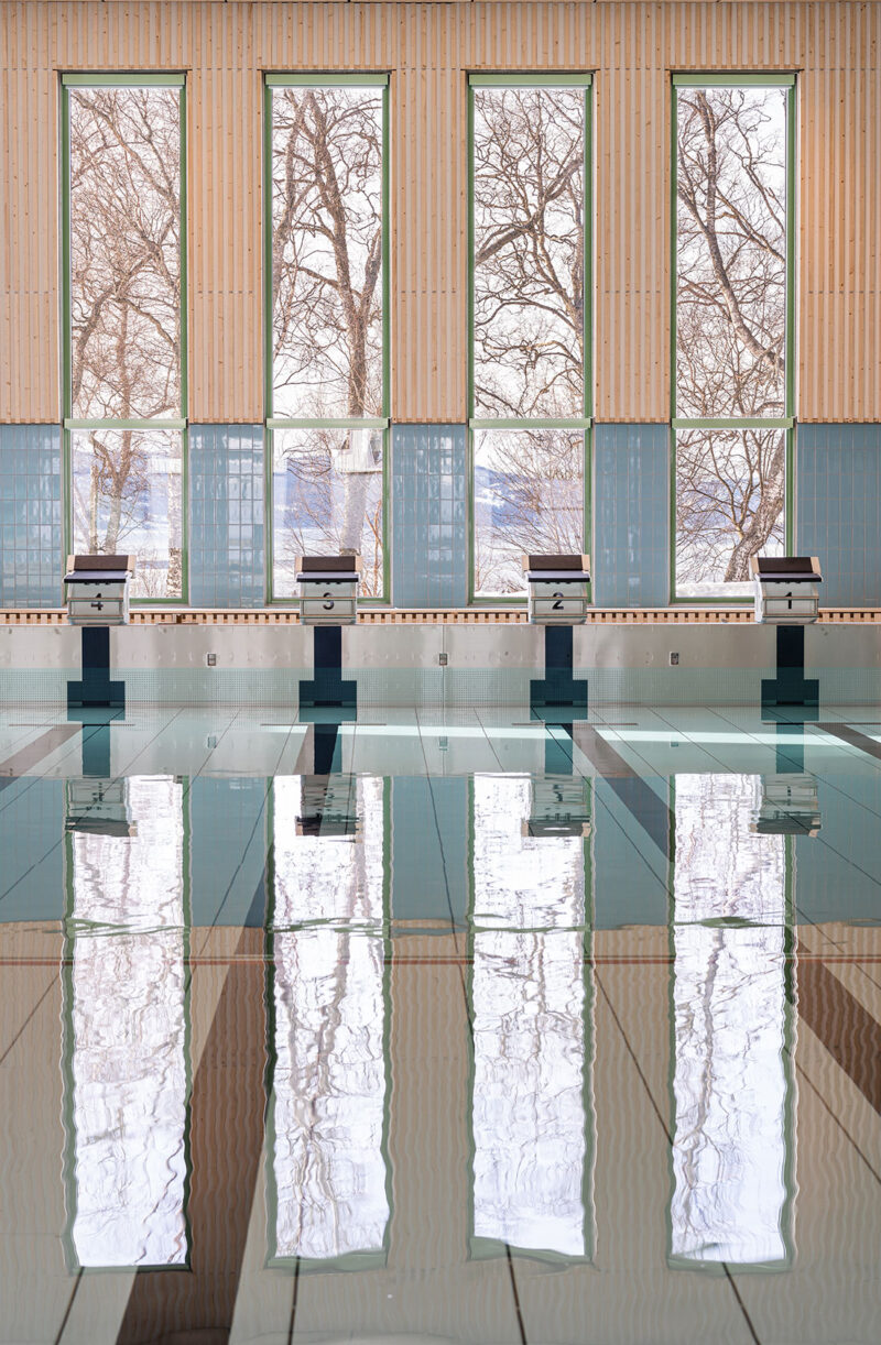 Indoor swimming pool with numbered starting blocks and large windows reflecting trees on the water surface.