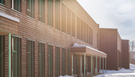 Modern wooden building in snowy landscape with sunlight filtering through, multiple windows visible.