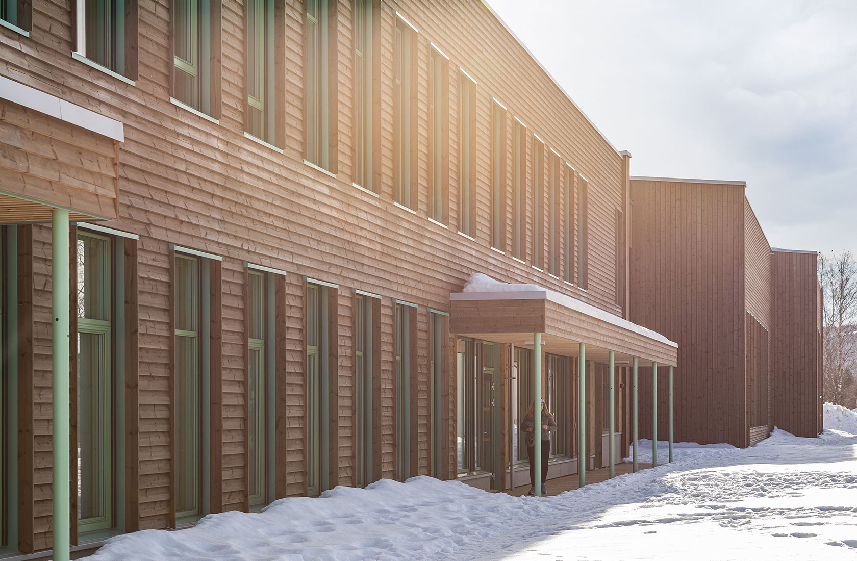 Modern wooden building in snowy landscape with sunlight filtering through, multiple windows visible.