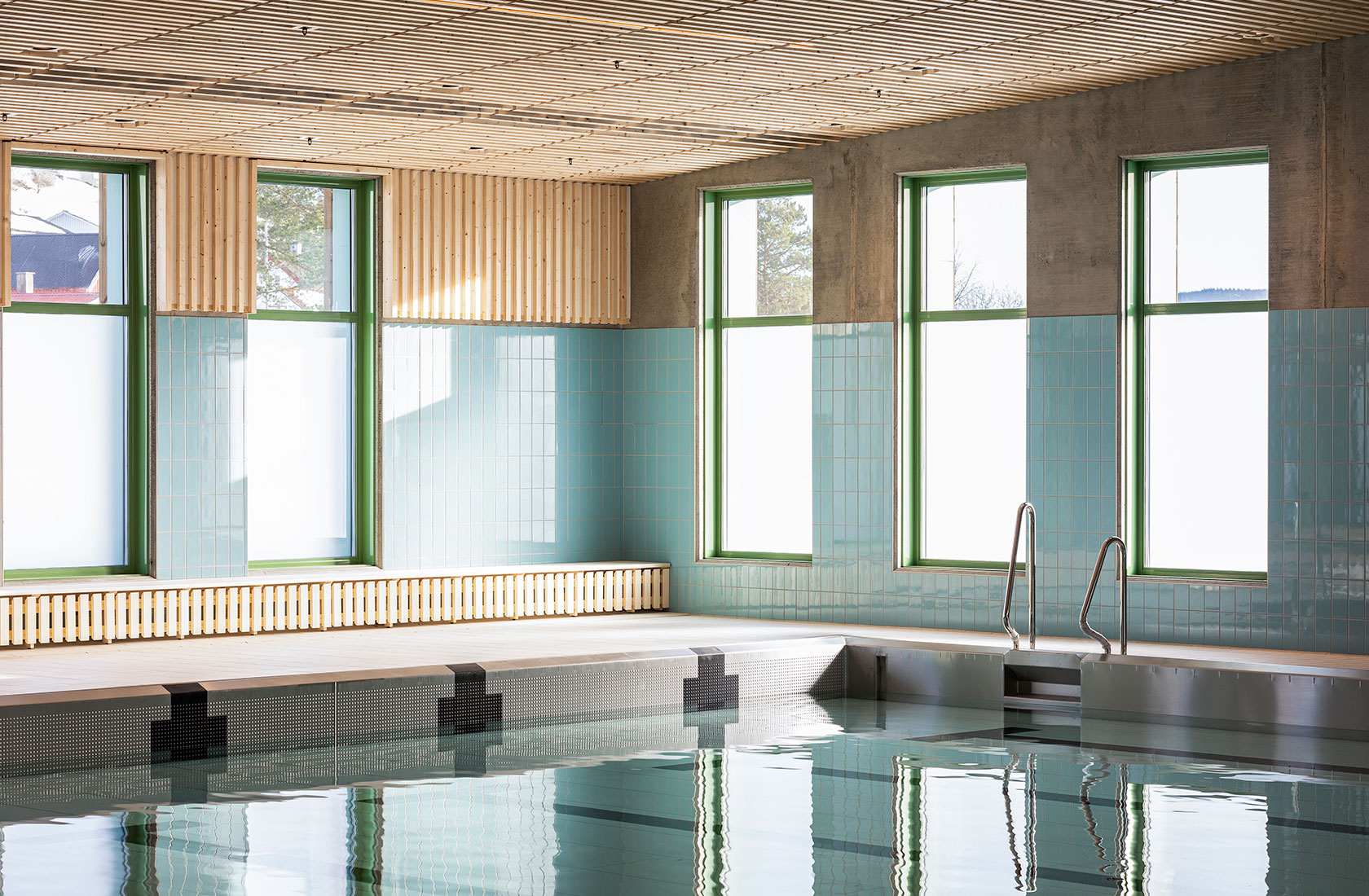 Indoor swimming pool with light blue tiles, large windows, and wooden ceiling.