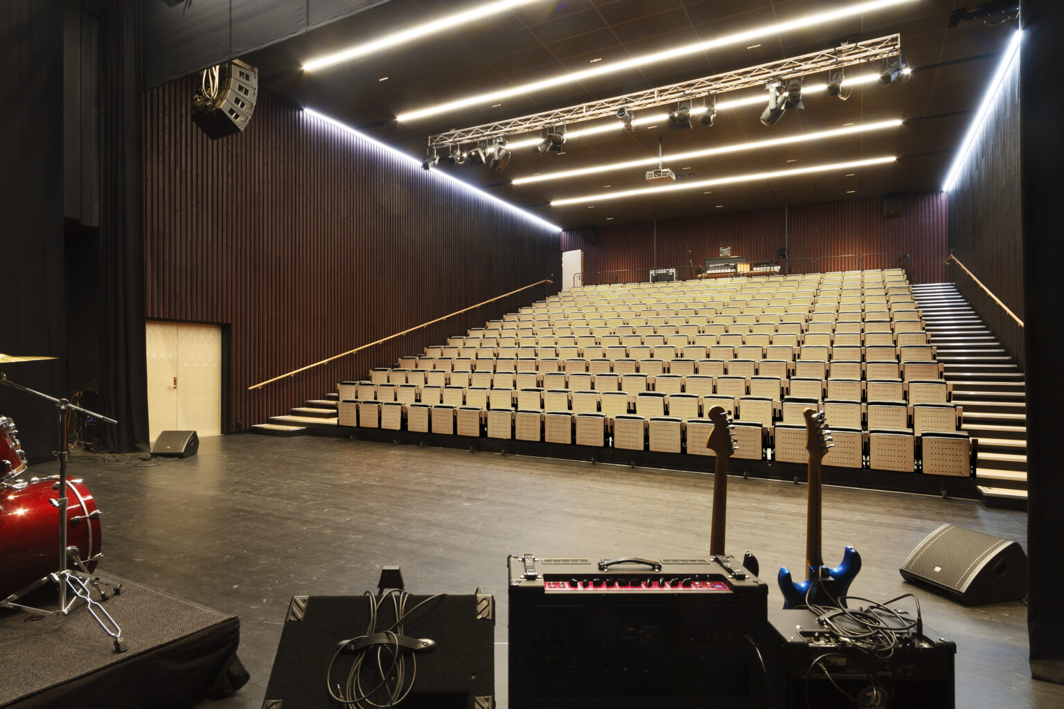 Empty concert hall with stage equipment, guitars, and drum set, rows of beige seats and modern lighting.