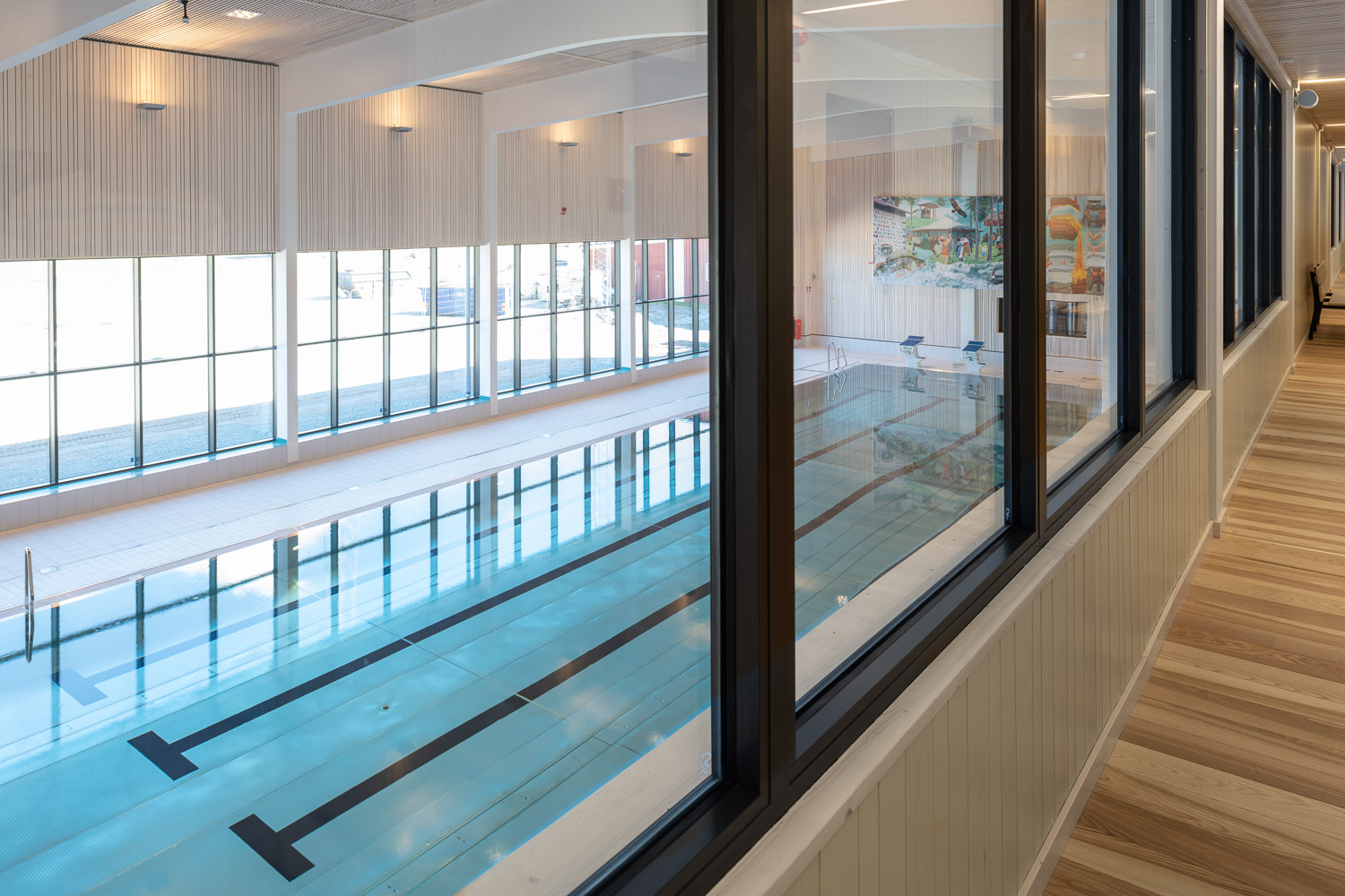 Indoor swimming pool with large windows and wooden floors, viewed from a glass-walled corridor.
