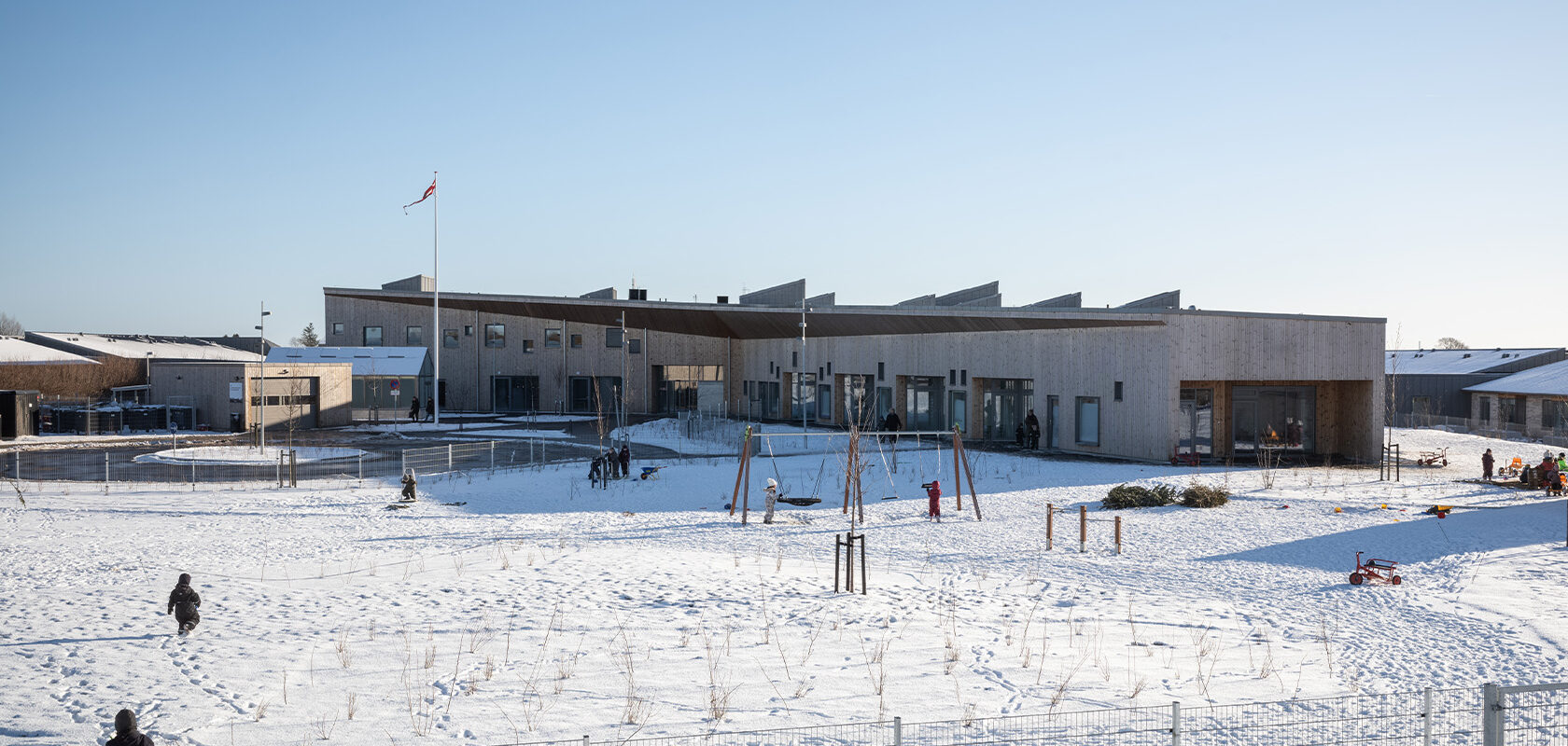 Modern wooden building with large windows surrounded by snow, children playing in the open area.