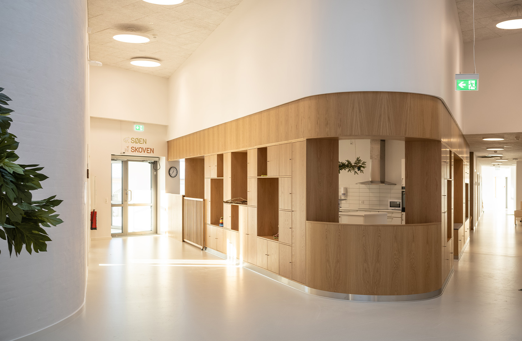 Modern indoor hallway with wooden cabinets, white walls, and ceiling lights. Doorway labeled "Søen Skoven."
