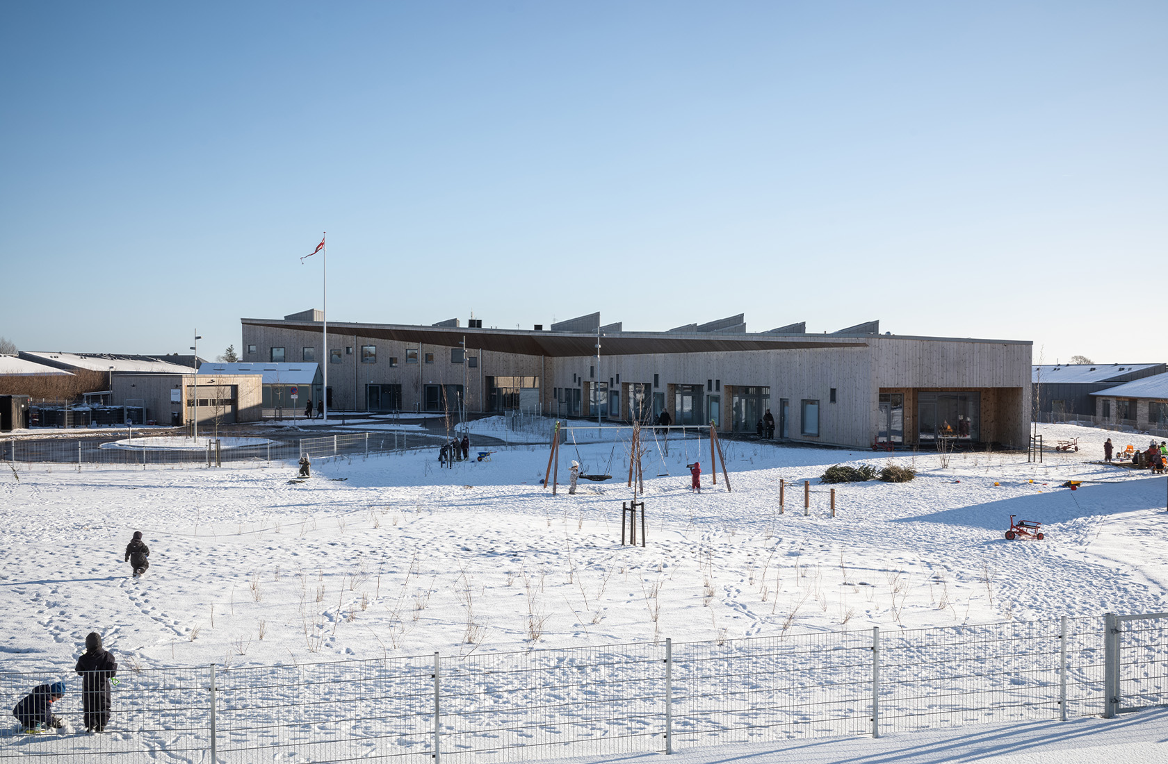 Modern wooden building with large windows surrounded by snow, children playing in the open area.