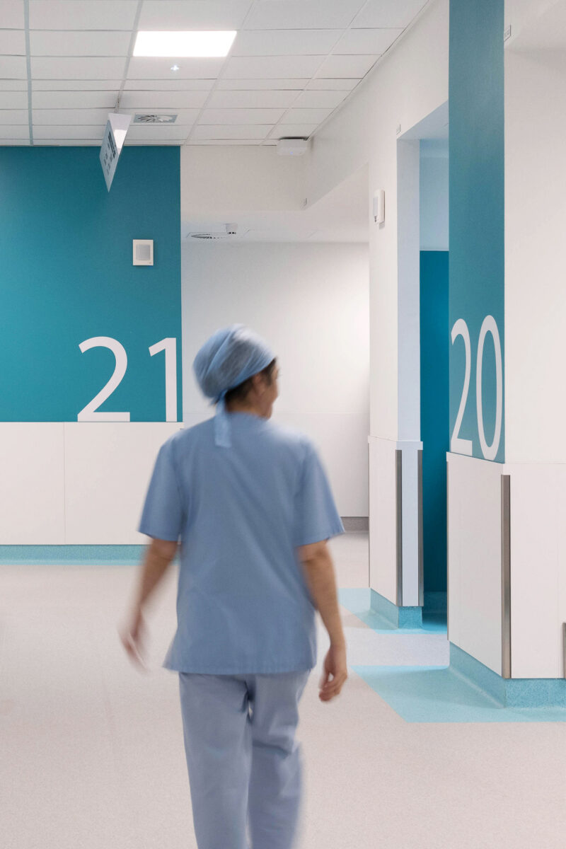 Hospital corridor with nurse in scrubs walking past room numbers 20 and 21