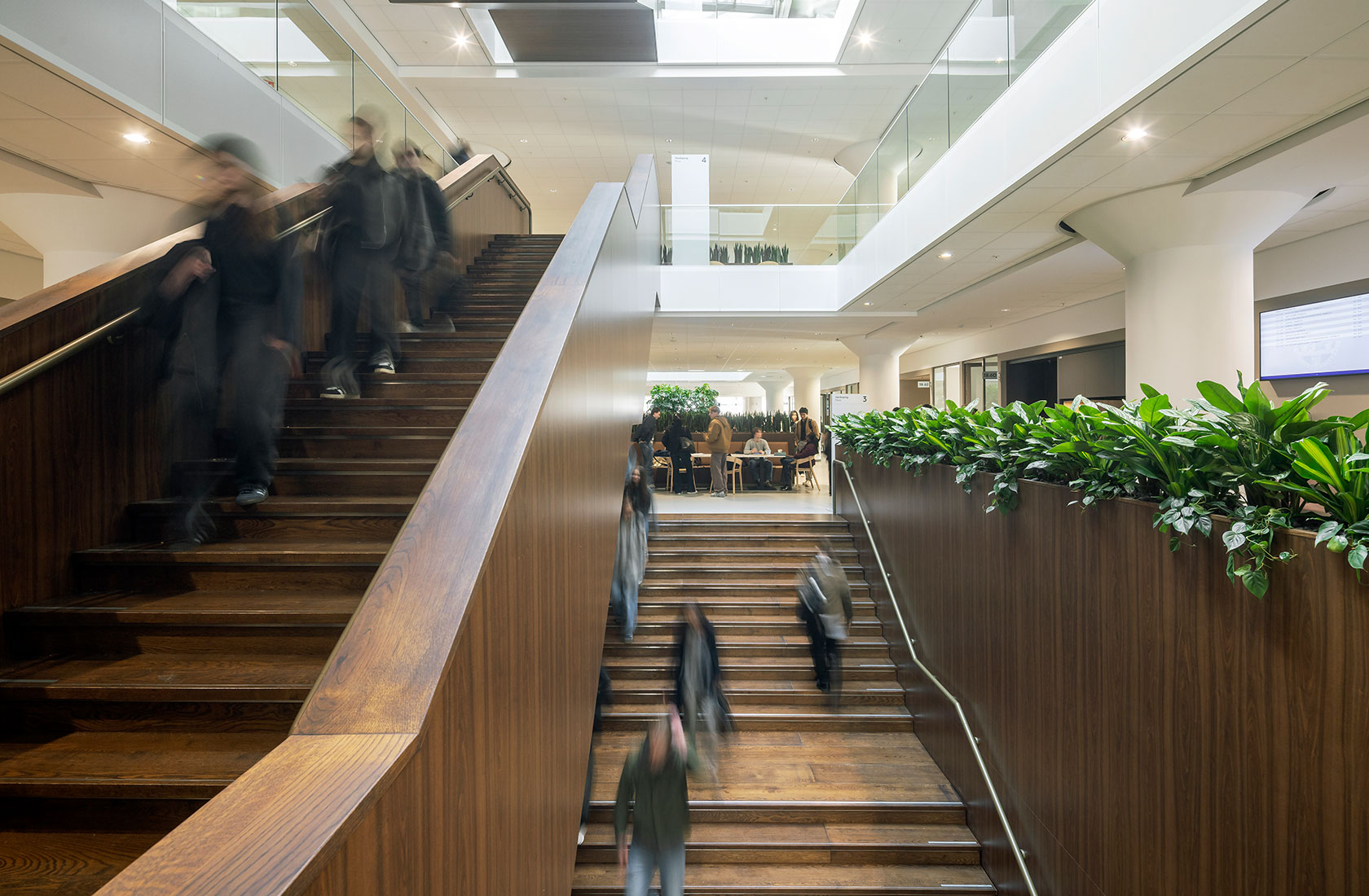 Modern university atrium with wooden staircase, blurred people walking, and indoor plants