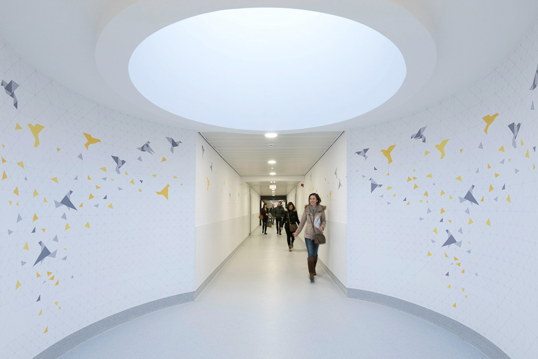 Modern hospital hallway with geometric wall art and people walking under circular skylight