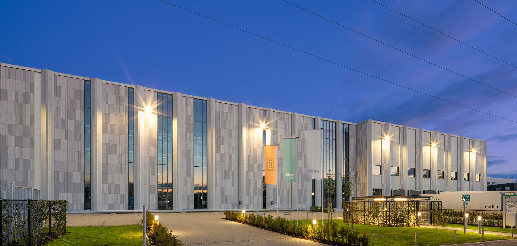 Modern industrial building exterior at dusk with lit facade and landscaped entrance walkway