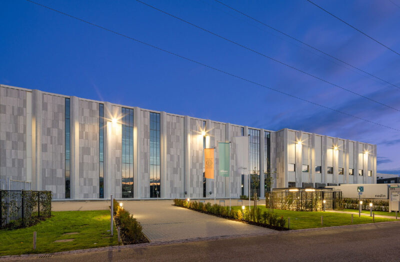 Modern industrial building exterior at dusk with lit facade and landscaped entrance walkway