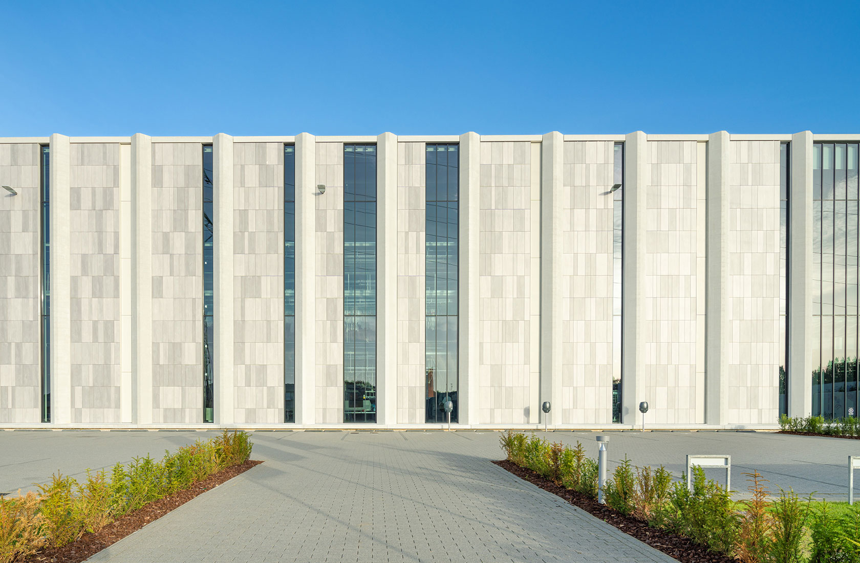Modern white concrete industrial building facade with tall glass windows under clear blue sky.