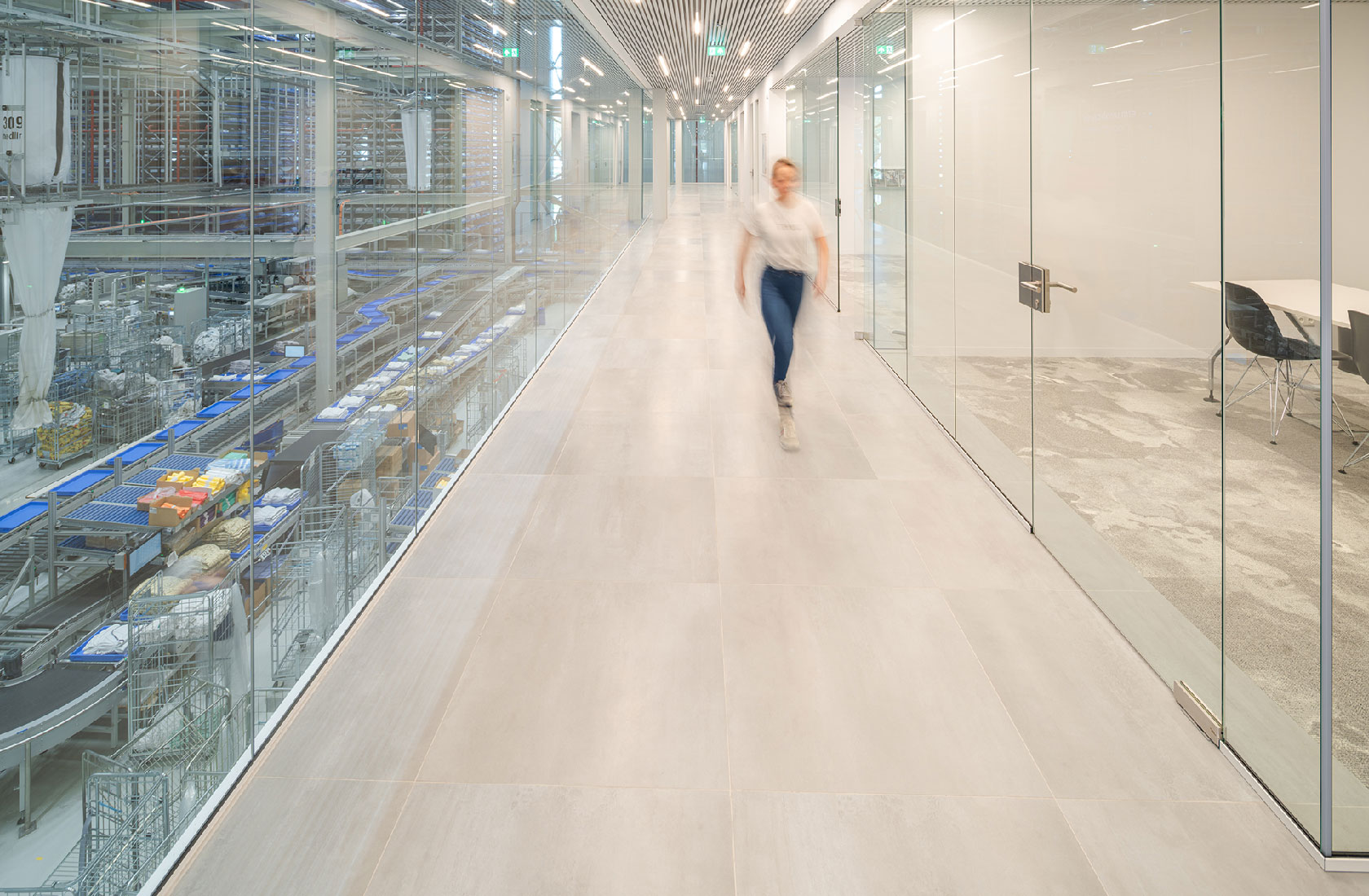 Modern industrial building corridor with glass walls overlooking automated warehouse conveyors.